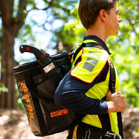 A man wearing a high-visibility safety vest carries a large black Reecoil Rope Bags backpack outdoors in a wooded area.