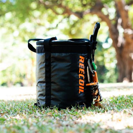 Black and orange REECOIL rope bag on grass with blurred trees in the background