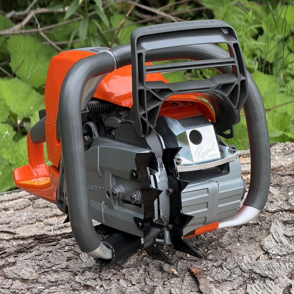 A Ripsaw RS572 - A ported Husqvarna chainsaw on a log with foliage in the background. The saw is shown with Ripsaw 4 point felling dogs and a Ripsaw Signature Series Exhaust.